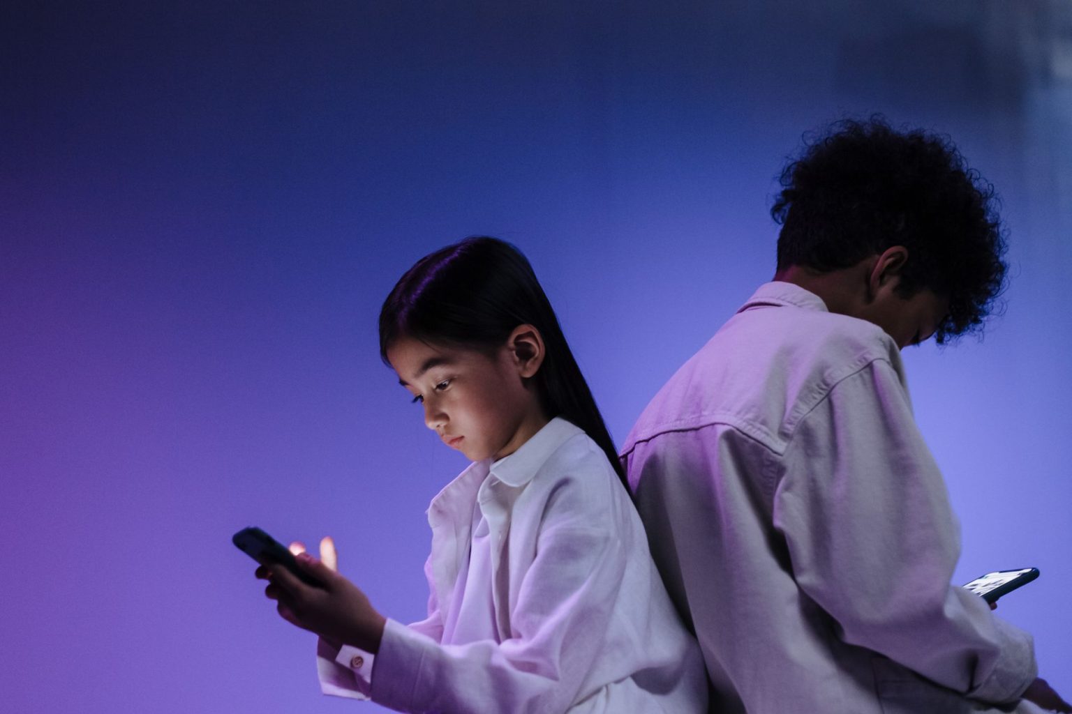 Duas crianças usam smartphones de costas uma para a outra sob iluminação colorida.