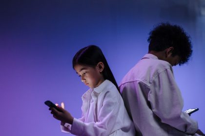 Duas crianças usam smartphones de costas uma para a outra sob iluminação colorida.