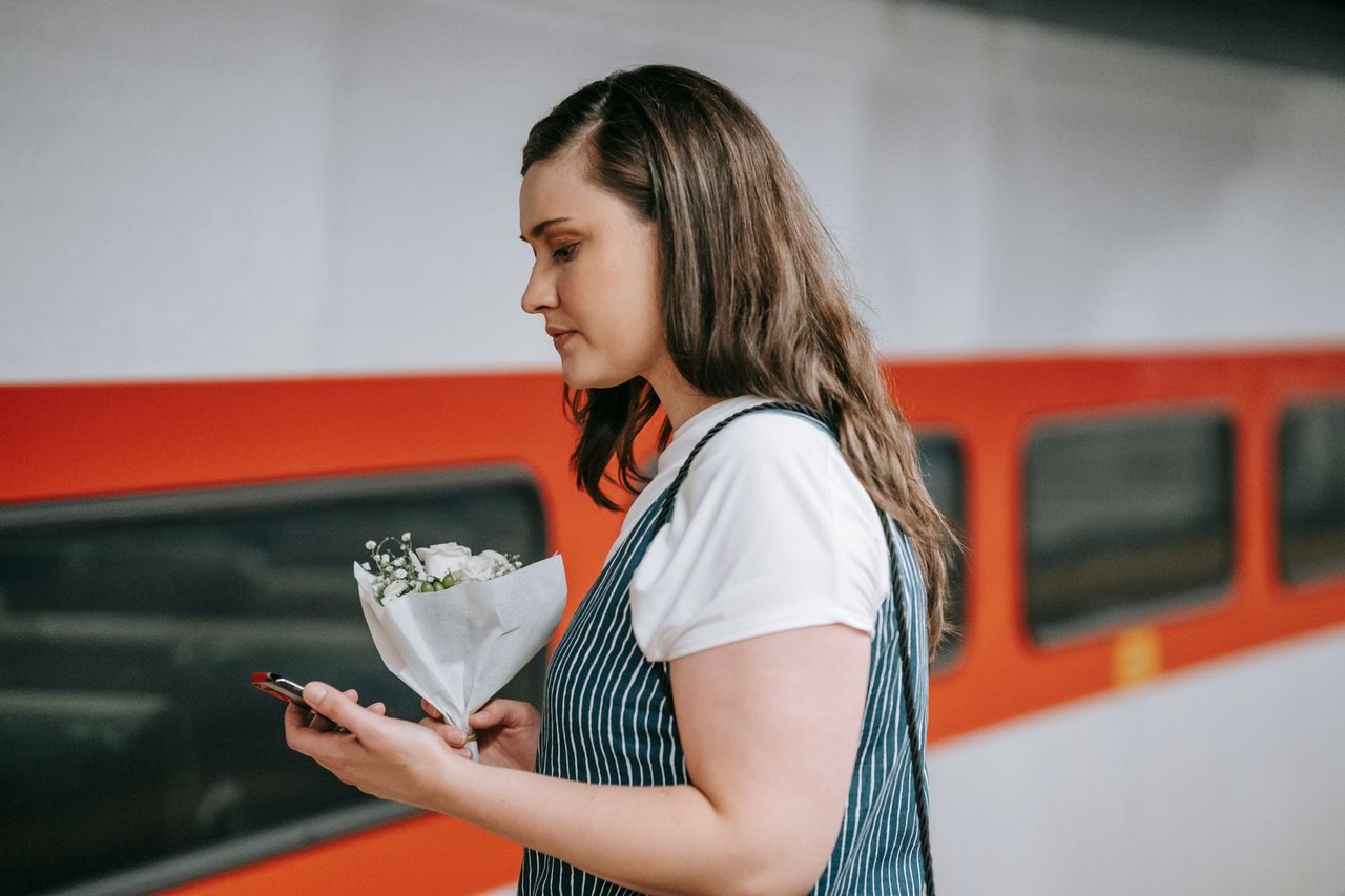 Passageira usa o celular em uma estação de trem enquanto segura flores.