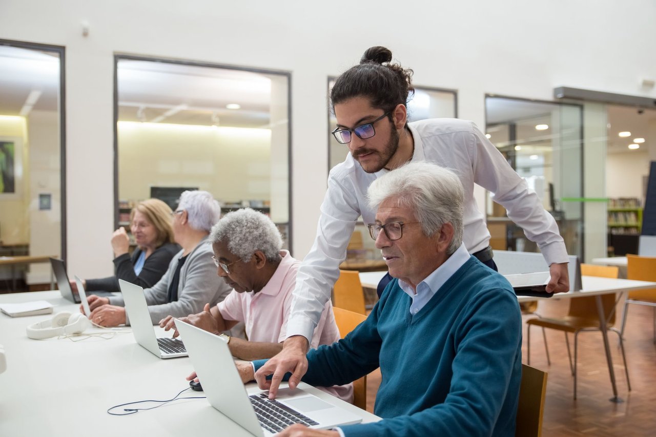 Professor orienta idosos que usam laptops durante uma atividade de aprendizagem em uma biblioteca.